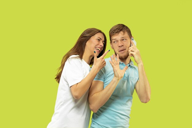 Beautiful young couple's half-length portrait isolated. Man speaking on the phone, woman is angry for it. Facial expression, human emotions concept. Trendy colors.
