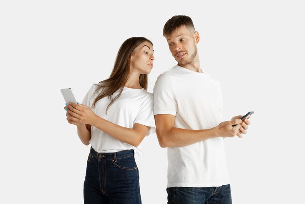 Beautiful young couple's portrait isolated on white studio background. Facial expression, human emotions, advertising concept. Both holding smartphones, woman watching in his screen. Keeping secrets.