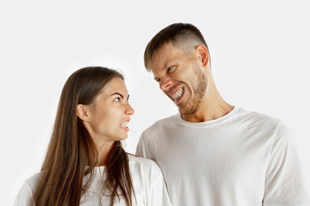 Beautiful young couple's portrait isolated on white wall. Facial expression, human emotions, advertising concept. Man and woman standing, looking at each other with an angry.