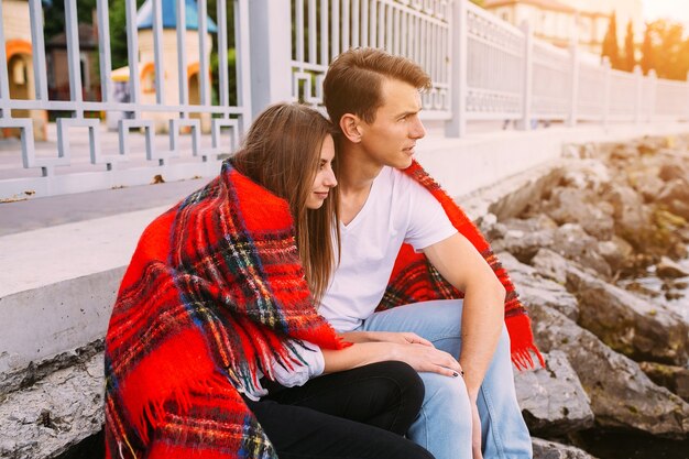 Beautiful young couple sitting on a stone embankment, wrapped in a blanket