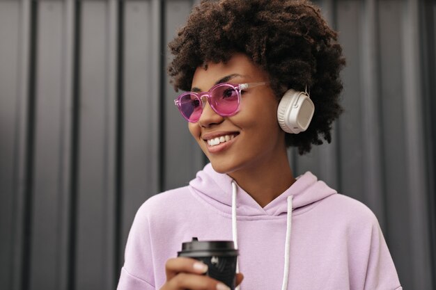 パステルで音楽を聴いている陽気な黒人の女の子 ヘッドフォンでポーズをとる紫色のセーターを着た魅力的なアフリカの女性 無料の写真
