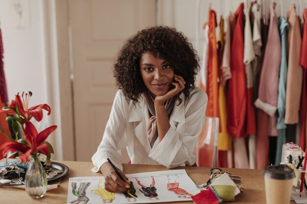 Beautiful young dark-skinned curly brunette woman in white blouse looks at front, leans on table and designs stylish clothes