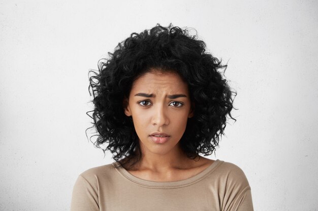 Beautiful young dark-skinned woman wearing beige top frowning looking, having clueless facial expression, feeling confused