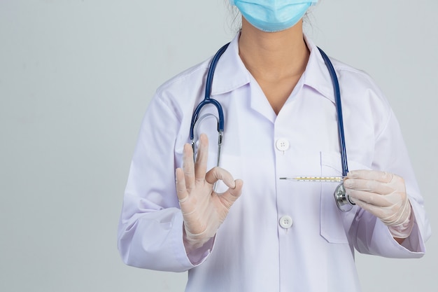 Beautiful young doctor is wearing mask while holding thermometer with rubber gloves on gray wall.