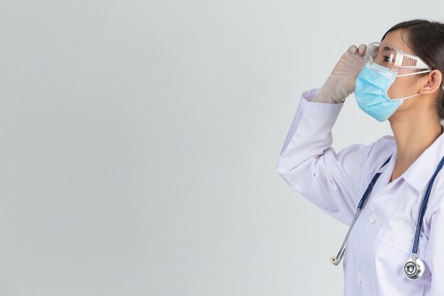 Beautiful young doctor is wearing mask while touch her glasses on with rubber gloves on gray wall.