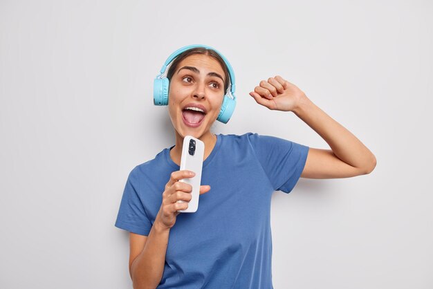 Beautiful young European woman dances and sing song holds modern smartphone as if microphone uses wireless stereo headphones wears casual t shirt has upbeat mood isolated over white background