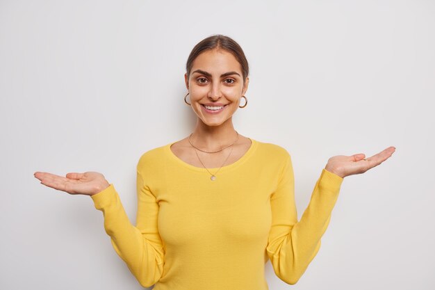 Beautiful young European woman smiles gently raises palms spreads hands over white wall demonstrates something wears casual yellow jumper pretends holding something
