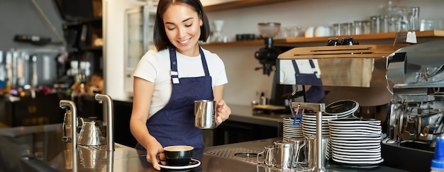 美しい若い女性のバリスタがカプチーノを作り ラテ・アートのための蒸しミルクをカップに注ぐ