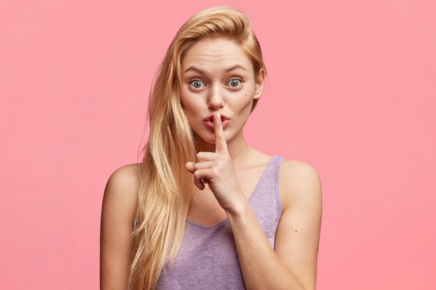 Beautiful young female blonde young woman keeps fore finger on lips, tells confidential information, demands complete silence