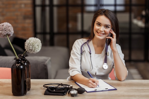 Beautiful young female medical and private doctor is looking at camera and smiling while give a recipe during call.