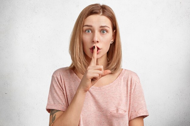 beautiful young female model asks to be quiet people who surround her, looks with warm blue eyes at camera, keeps fore fingers on lips
