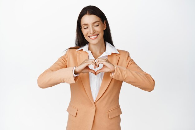 Beautiful young female office manager close eyes and shows heart sign smiling carefree standing in suit against white background Copy space