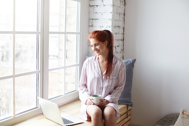 Beautiful young female with cute smile and ginger hair in ponytail enjoying slow morning at home, sitting by large window, drinking coffee, surfing internet on laptop. Daylight sunshine, soft colors