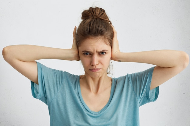 Foto gratuita bella giovane donna con il nodo dei capelli che ha uno sguardo scontento che si tappi le orecchie infastidita dal rumore che vuole il silenzio e l'atmosfera calma essendo esausta di suoni forti e gridando intorno a lei