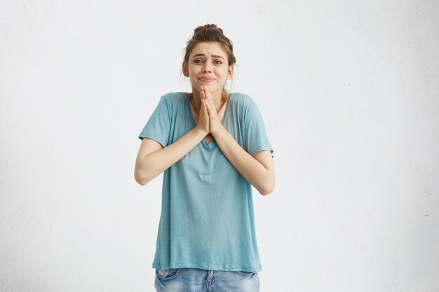 Beautiful young female with imploring appealing look holding palms pressed together in front of her, asking for forgiveness, feeling sorry