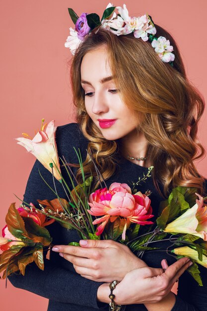 Красивая молодая женщина с длинными волнистыми светлыми волосами в венке из весенних цветов позирует с букетом цветов на розовом фоне.