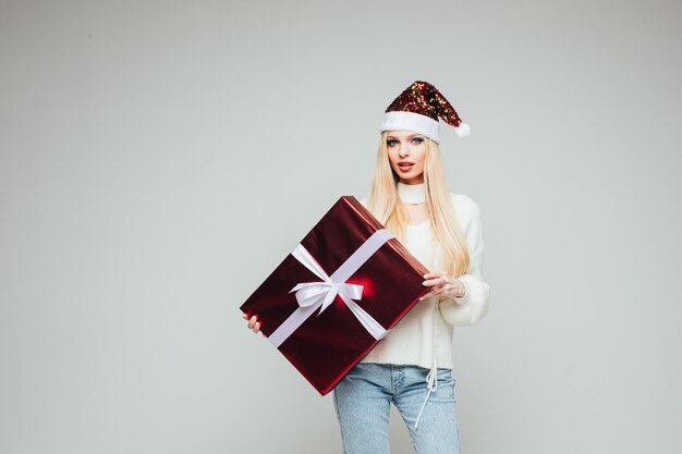 赤​と​白​の​クリスマス​の​帽子​を​持つ​美しい​若い​女性​は​、​プレゼント​と​喜び​の​大きな​箱​を​保持します