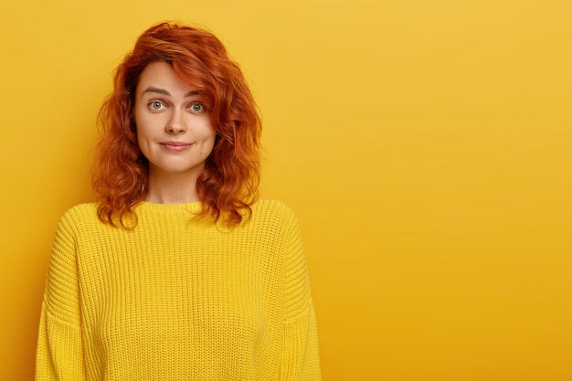 Beautiful young ginger woman wears bright knitted yellow sweater, raises eyebrows with wonder, looks surprisingly at camera
