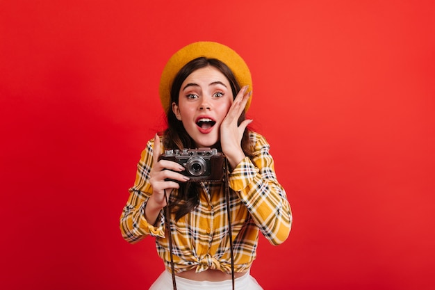 美しい少女ブロガーはレトロなカメラで写真を作ります。オレンジ色の衣装と赤い壁に帽子の緑色の目の女性の肖像画。