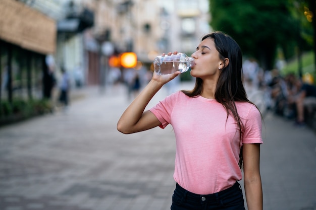 Красивая молодая девушка пьет бутылку воды на городской улице