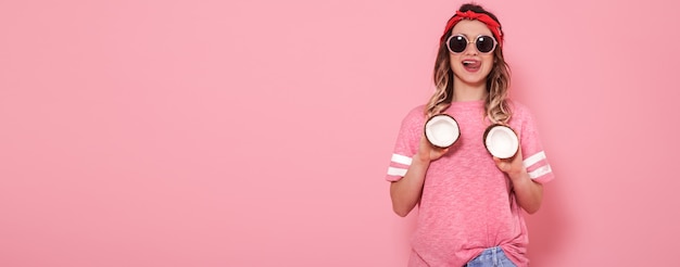 Beautiful young girl in pink t-shirt and glasses holding funny sexy coconuts on pink background