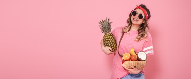 Красивая молодая девушка в розовой футболке и очках, держит полную соломенную сумку с фруктами на розовом фоне