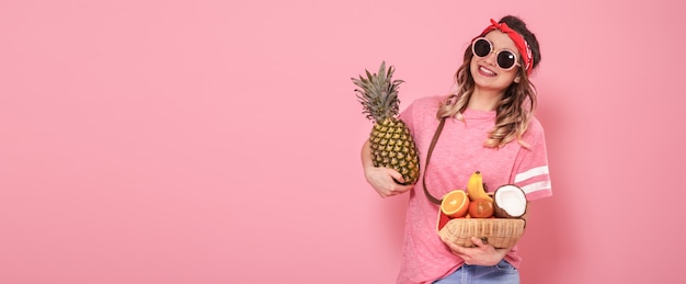 ピンクのtシャツとメガネの美しい少女は、ピンクの背景に果物の完全なストローバッグを保持しています。