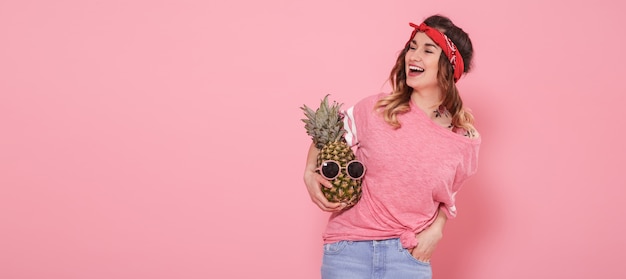 Beautiful young girl in pink t-shirt, smiling with pineapple on pink wall