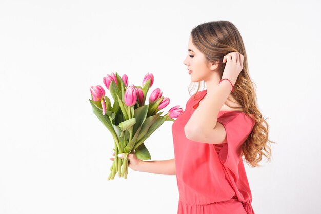 A beautiful young girl stands on a white wall, wearing a pink dress, and a bouquet of tulips