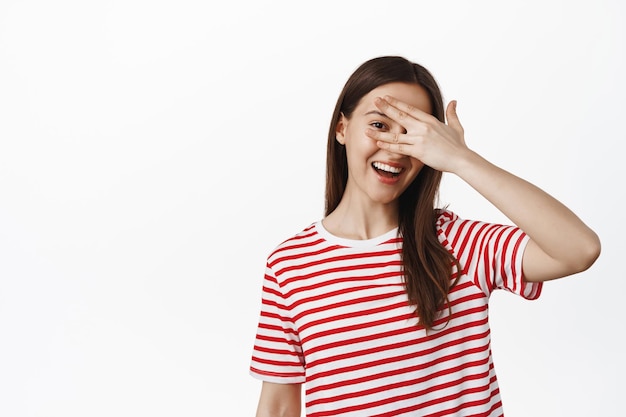 Beautiful young girl student, wearing t-shirt, open one eye, peeking through fingers and smiling at you, looking at smth, standing against white background.