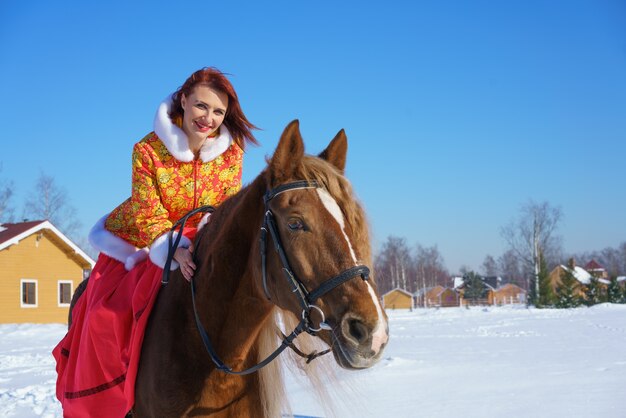 따뜻한 노란색-빨간색 재킷을 입은 아름다운 어린 소녀가 겨울 맑은 서리가 내린 날에 말을 탄다. 겨울철에 승마 스포츠에 종사