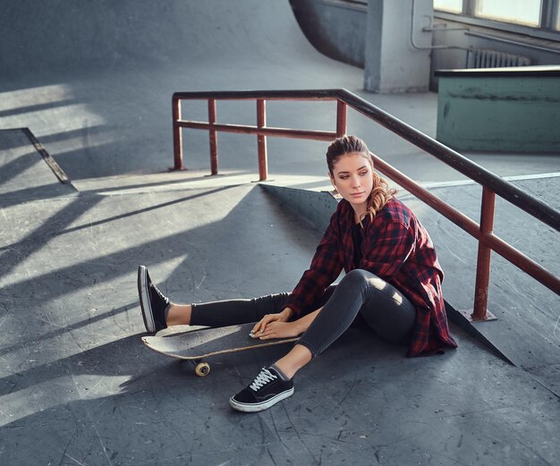 A beautiful young girl wearing a checkered shirt holding a skateboard while sitting next to a grind rail in skatepark indoors.