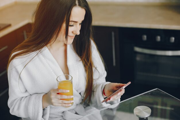 beautiful young girl with black hair and white robe sitting at home in the kitchen at the table 