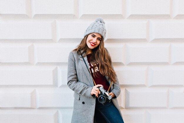 Beautiful young girl with long hair in grey coat and knitted hat on grey wall . She holds camera in hands and looks enjoyed.