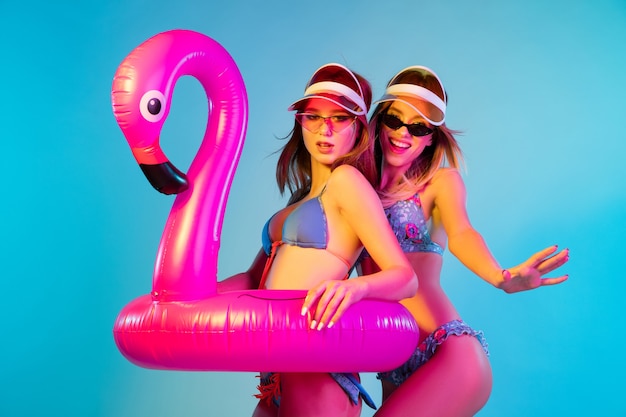 Beautiful young girls' half-length portrait isolated on blue  wall in neon light. Women posing in fashionable bodysuit. Facial expression, summer, weekend concept. Trendy colors.