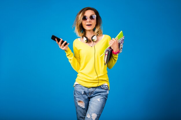 Beautiful young hipster woman holding books and looking at smartphone