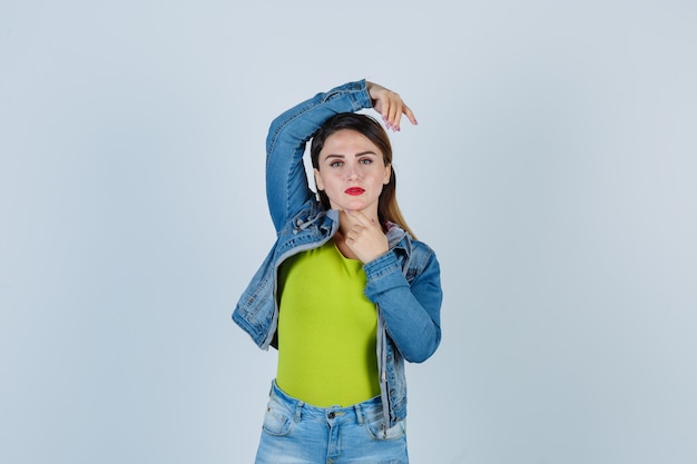 Beautiful young lady in denim outfit posing with hands around head and looking alluring , front view.