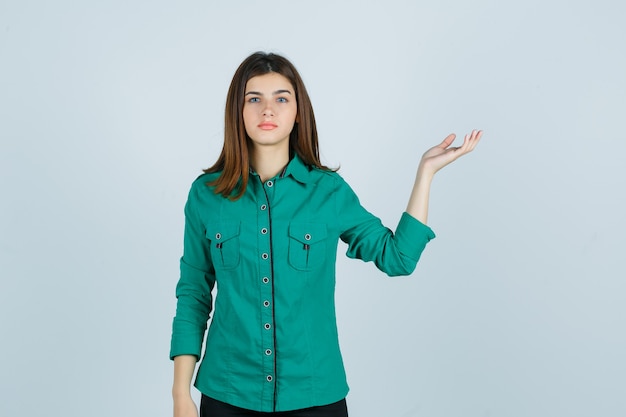 緑のシャツでウェルカムジェスチャーをし、困惑した、正面図を見て美しい若い女性。