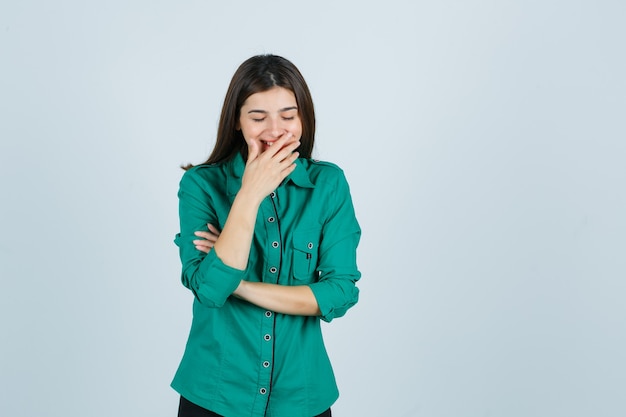 Bella giovane signora in camicia verde che tiene la mano sulla bocca e che sembra felice, vista frontale.