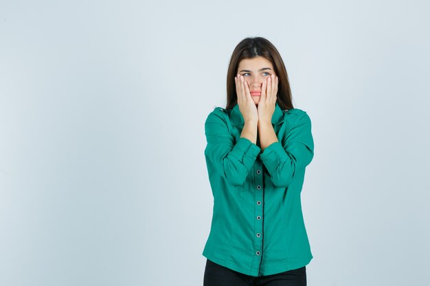 Beautiful young lady holding hands on cheeks in green shirt and looking downcast , front view.