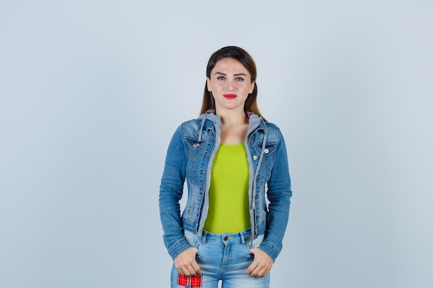 Beautiful young lady keeping thumbs in pockets in denim outfit and looking cheerful , front view.