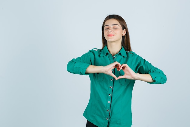 Beautiful young lady making heart gesture in green shirt and looking peaceful , front view.