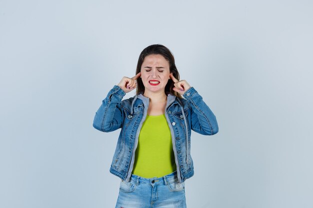 Beautiful young lady plugging ears with fingers in denim outfit and looking annoyed. front view.