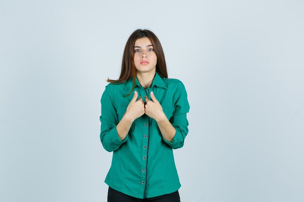 Beautiful young lady pointing at herself in green shirt and looking bewildered , front view.