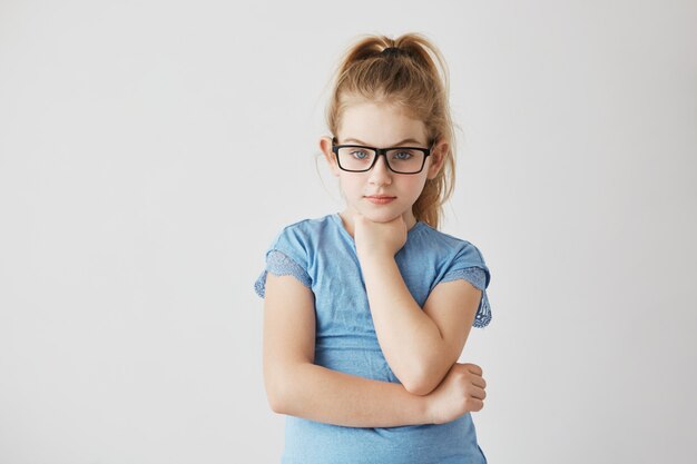 Free photo beautiful young lady with bright blue eyes and light hair posing in glasses with hand under her chin and serious face expression.