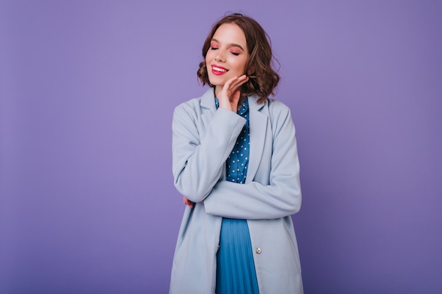 Красивая барышня с ярким макияжем позирует в осенней одежде. Крытая фотография очаровательной кудрявой девушки в синем пальто, изолированном на фиолетовой стене.
