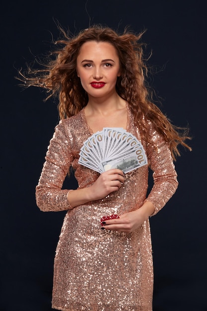 Free photo beautiful young lucky happy brown-haired woman in golden cocktail dress showing the money fan in one hand and poker chips in another hand, fan of 100 dollar bills, black background. gambling, casino,