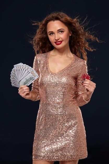 Free photo beautiful young lucky happy brown-haired woman in golden cocktail dress showing the money fan in one hand and poker chips in another hand, fan of 100 dollar bills, black background. gambling, casino,