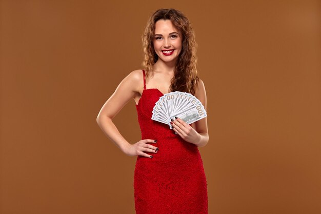 Красивая молодая счастливая шатенка в красном коктейльном платье показывает фанату денег 100-долларовые купюры, коричневый фон. Успех, удача, лотерея