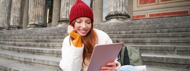 Foto gratuita la bella giovane ragazza moderna con capelli rossi tiene la compressa digitale si siede sulle scale vicino al museo e